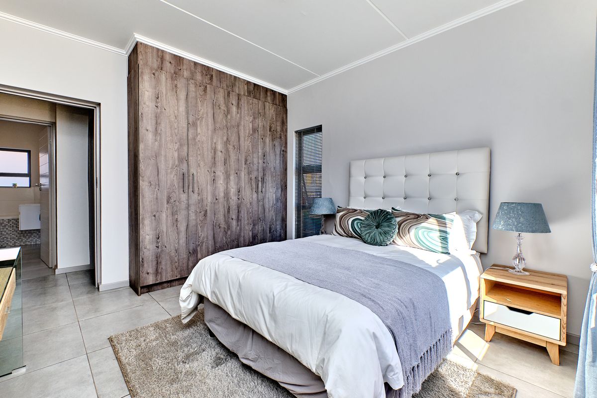 A well-maintained bedroom featuring modern wood-finish built-in cupboards, neutral tiled flooring, and a clean, contemporary aesthetic with access to an adjoining bathroom.