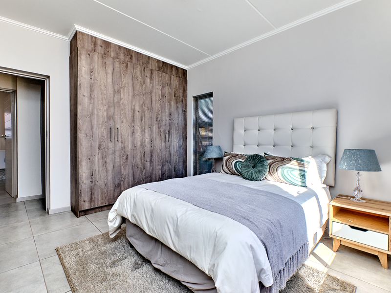 A well-maintained bedroom featuring modern wood-finish built-in cupboards, neutral tiled flooring, and a clean, contemporary aesthetic with access to an adjoining bathroom.