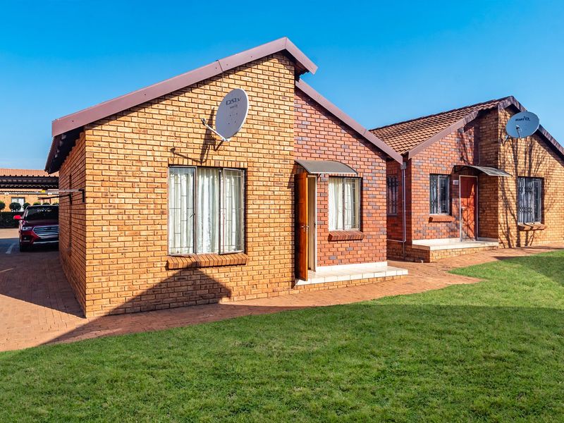 A well-maintained face brick townhouse exterior featuring a paved driveway, tiled roof, and satellite dish installation in a neat residential complex.