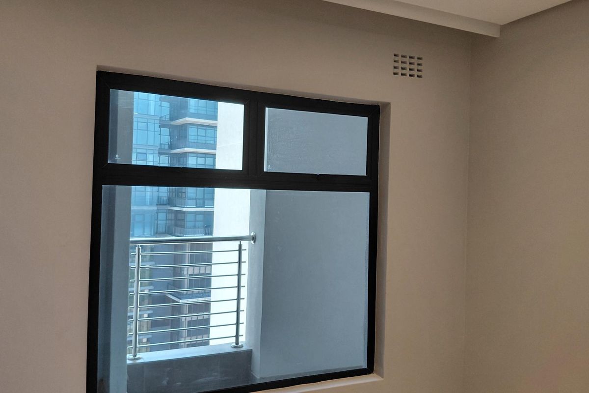 A bright bedroom featuring a large window with black frames, offering a view of the adjacent building and balcony.