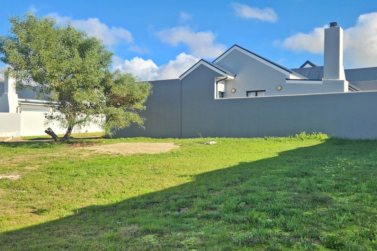 A view of a vacant residential plot featuring a grass lawn, boundary wall, and adjacent residential structure with a chimney.