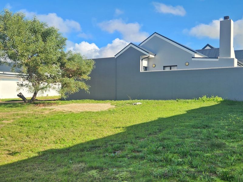 A view of a vacant residential plot featuring a grass lawn, boundary wall, and adjacent residential structure with a chimney.