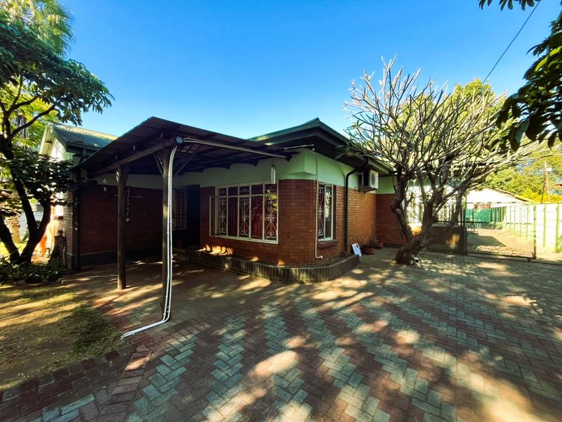 Front view of a brick house featuring a covered patio area, paved driveway, and established garden surroundings.