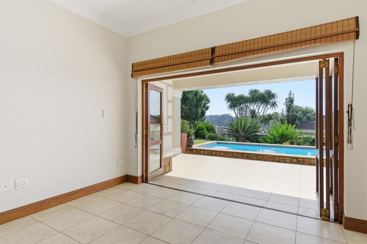 An interior living space featuring large wooden folding doors that open onto a tiled patio area with a swimming pool and garden views.