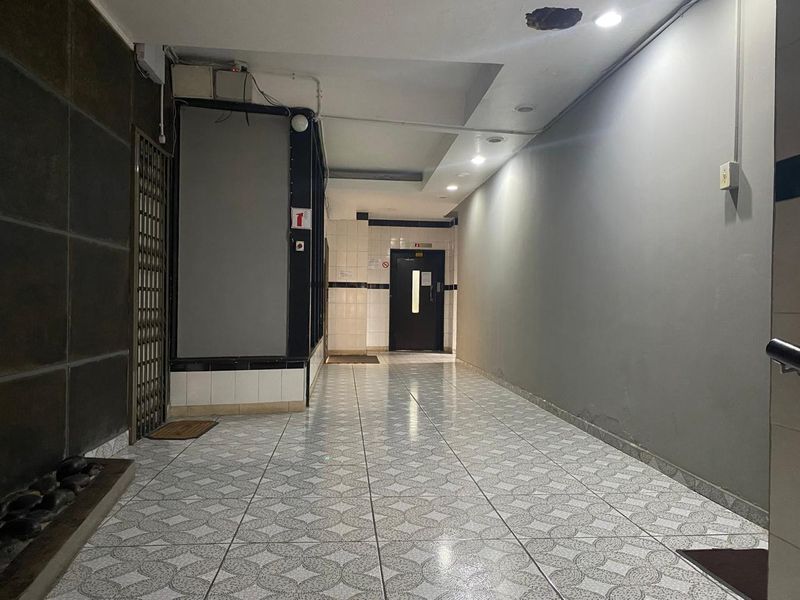 A communal apartment hallway featuring tiled flooring, recessed lighting, and access to an elevator, showing signs of wear and ceiling damage.