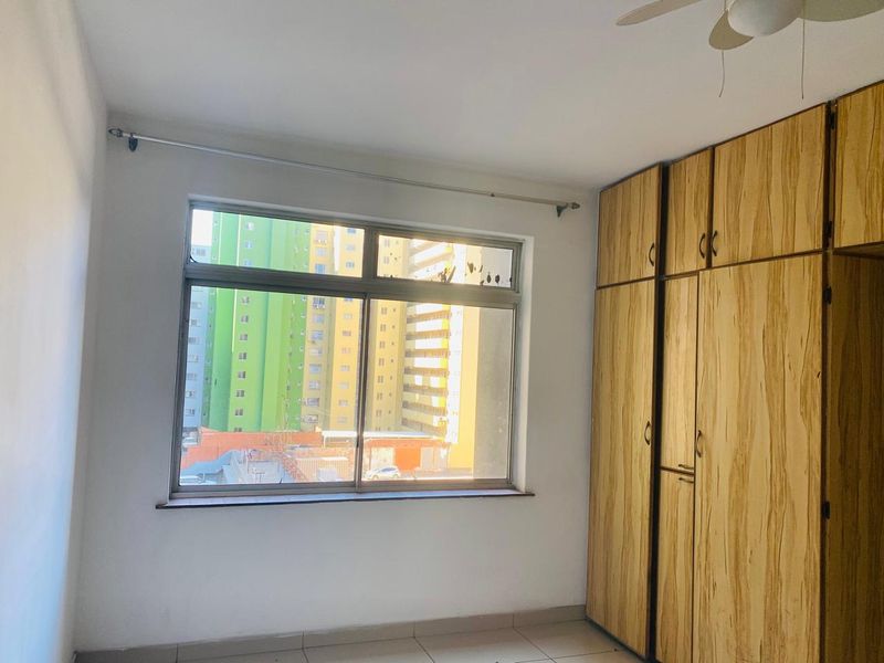 An empty bedroom featuring built-in wooden cupboards, tiled flooring, and an aluminium-framed window with an urban view.