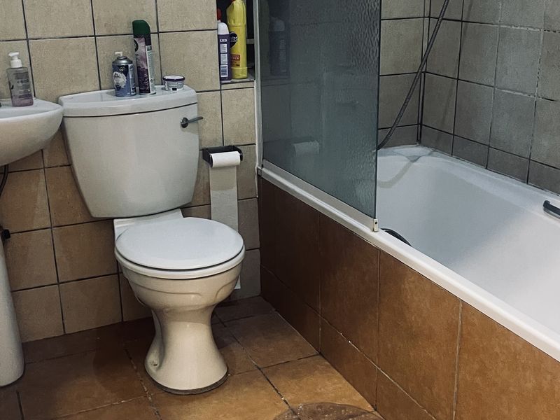A compact bathroom featuring a pedestal basin, toilet, and a bathtub with a glass shower screen, finished with dated ceramic wall and floor tiles.
