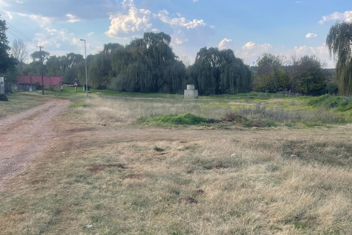 A view of a vacant residential plot featuring natural grass, mature trees, and a small concrete utility structure on the property.