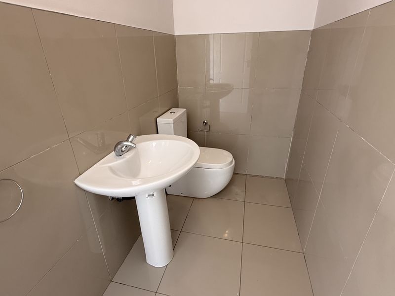 A clean, modern guest bathroom featuring neutral ceramic wall and floor tiles, a white pedestal washbasin, and a standard toilet.