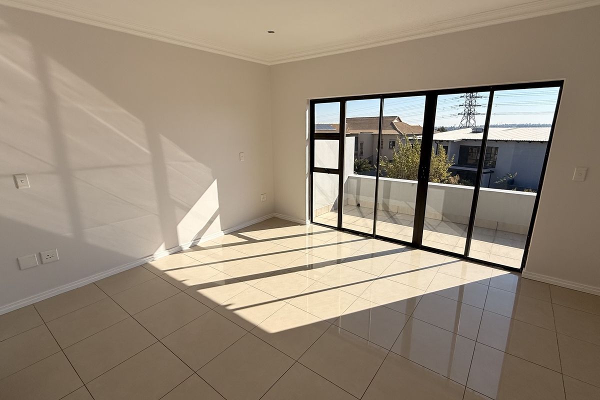 A bright, empty bedroom featuring large black aluminium sliding doors that open onto a private balcony, finished with neutral floor tiles.