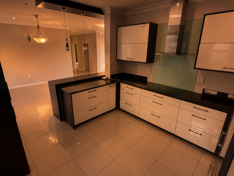 Modern open-plan kitchen featuring high-gloss cabinetry, granite countertops, and integrated appliances, seamlessly flowing into the adjacent living area with tiled flooring throughout.