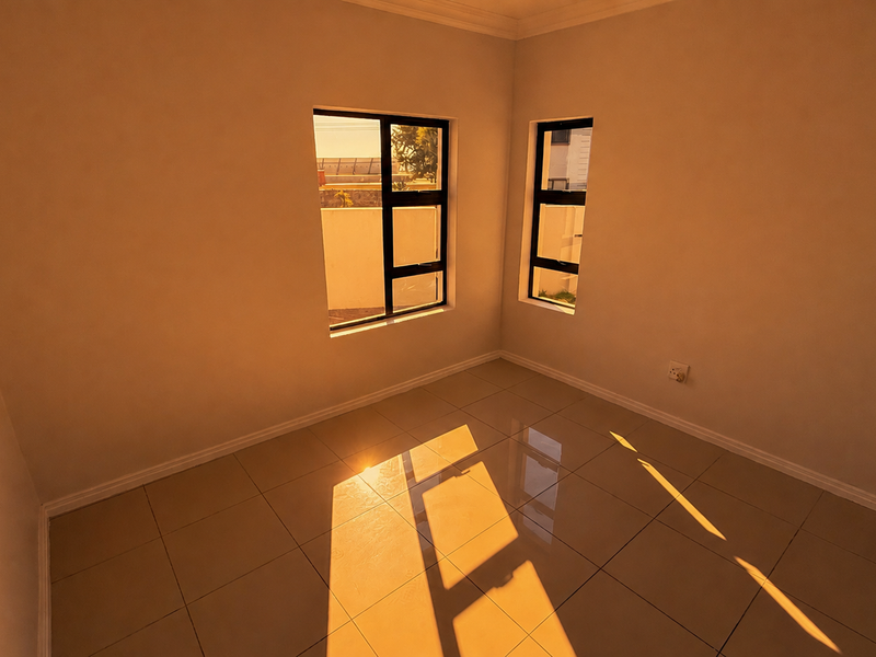 A bright, empty bedroom featuring neutral walls, polished floor tiles, and large aluminium-framed windows allowing for ample natural light.