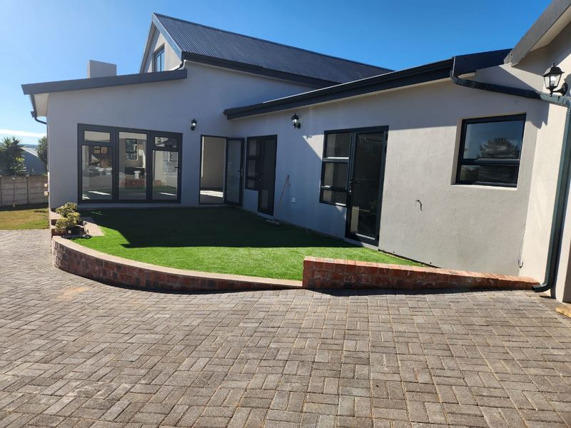 Rear exterior view featuring a paved driveway, brick retaining wall, artificial lawn, and modern aluminium-framed windows and doors.