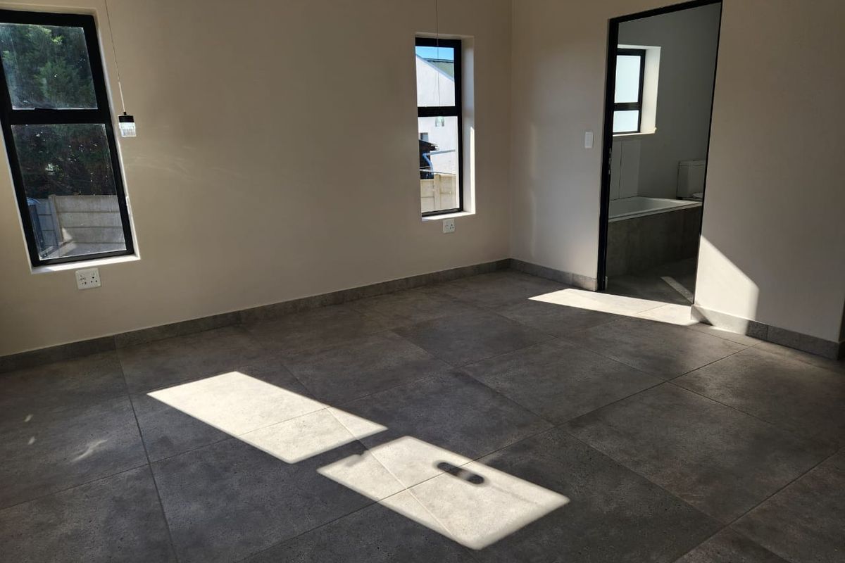 A bright, empty bedroom featuring modern grey floor tiles, aluminium window frames, and an adjoining en-suite bathroom.