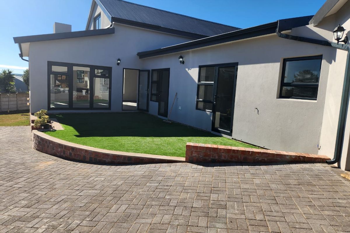 Rear exterior view featuring a paved driveway, brick retaining wall, artificial lawn, and modern aluminium-framed windows and doors.