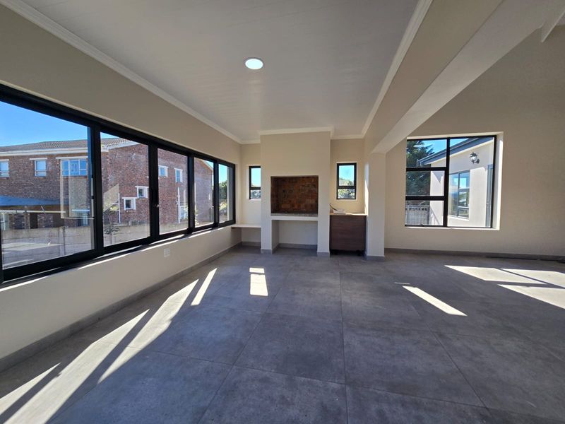 A bright, empty enclosed patio featuring large aluminium-framed windows, tiled flooring, and a built-in braai area, offering a versatile space for indoor-outdoor living.