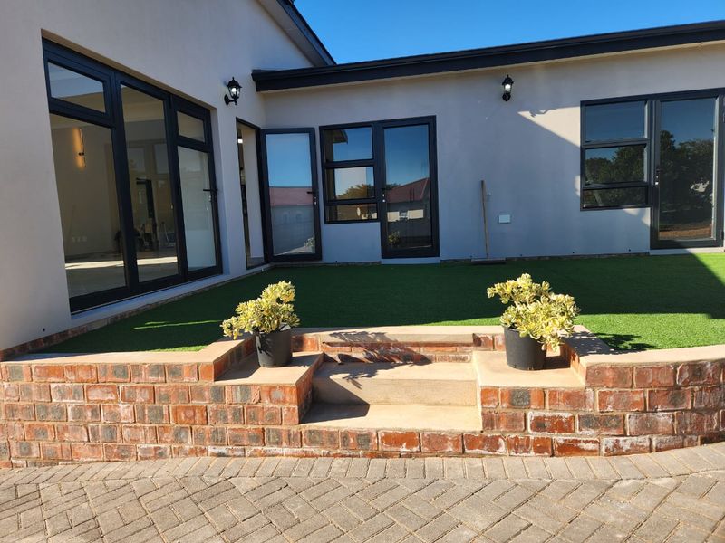 A well-maintained rear exterior featuring a brick retaining wall, concrete steps, artificial turf, and modern black aluminium doors and windows.