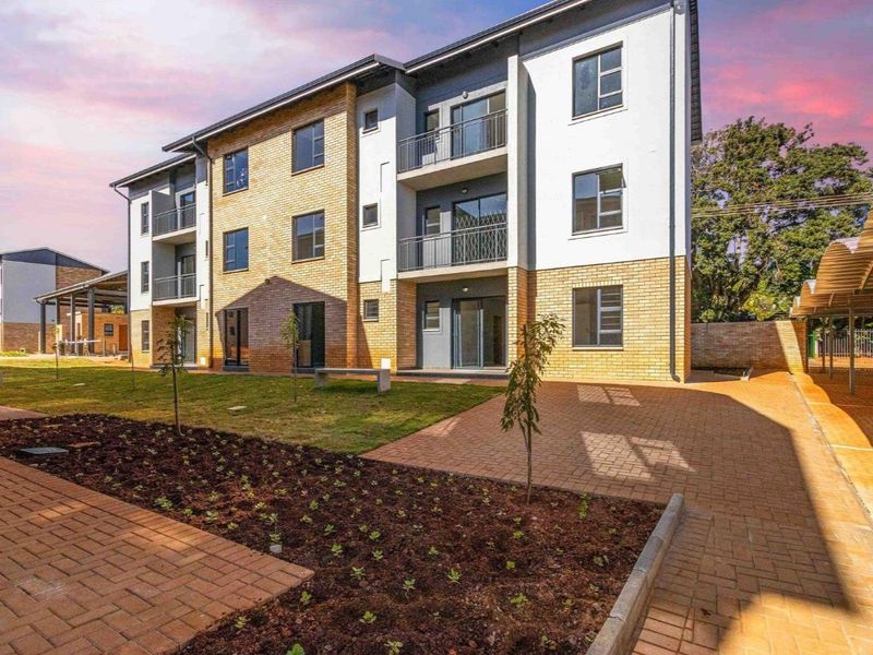 A modern multi-story apartment building featuring brick and plaster facade, private balconies, paved walkways, and landscaped garden areas.