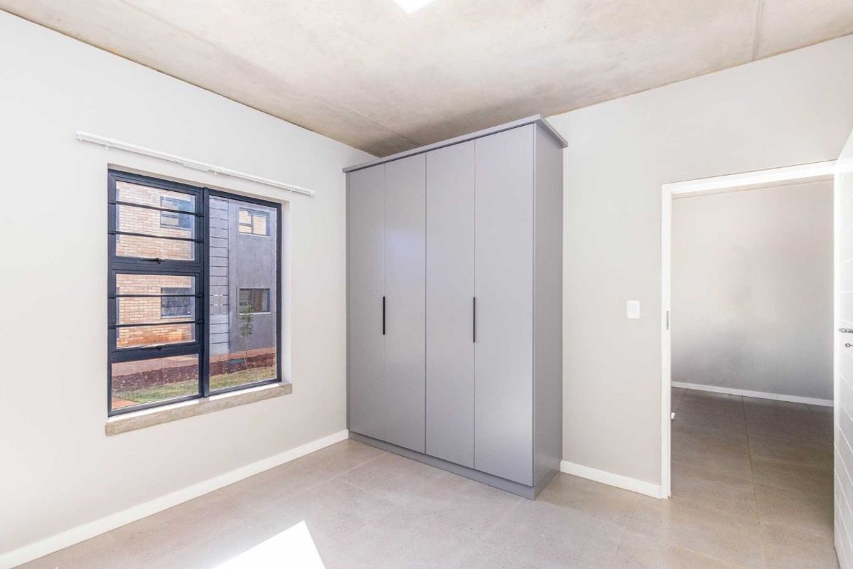 A bright, modern bedroom featuring neutral tiled flooring, an exposed concrete ceiling, and a sleek built-in wardrobe with grey cabinetry.