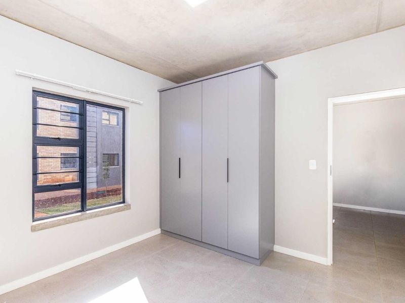 A bright, modern bedroom featuring neutral tiled flooring, an exposed concrete ceiling, and a sleek built-in wardrobe with grey cabinetry.