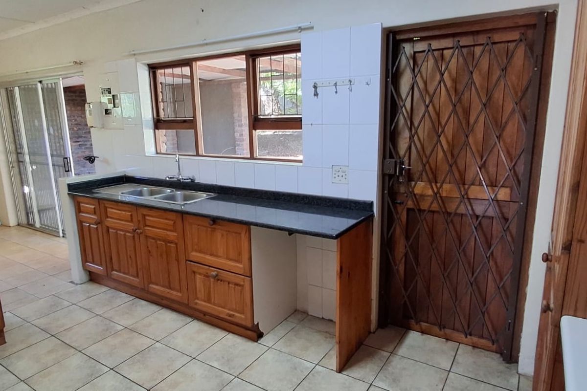 A functional kitchen featuring wooden cabinetry, granite countertops, a double basin sink, and tiled flooring, with direct access to an outdoor area.