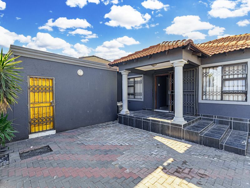 Front exterior view of the house featuring a paved driveway, a covered patio with concrete columns, and secure metal gates.