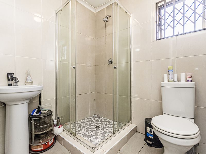 A functional bathroom featuring a pedestal basin, a glass-enclosed shower, and a standard toilet with neutral wall tiling and security bars on the window.