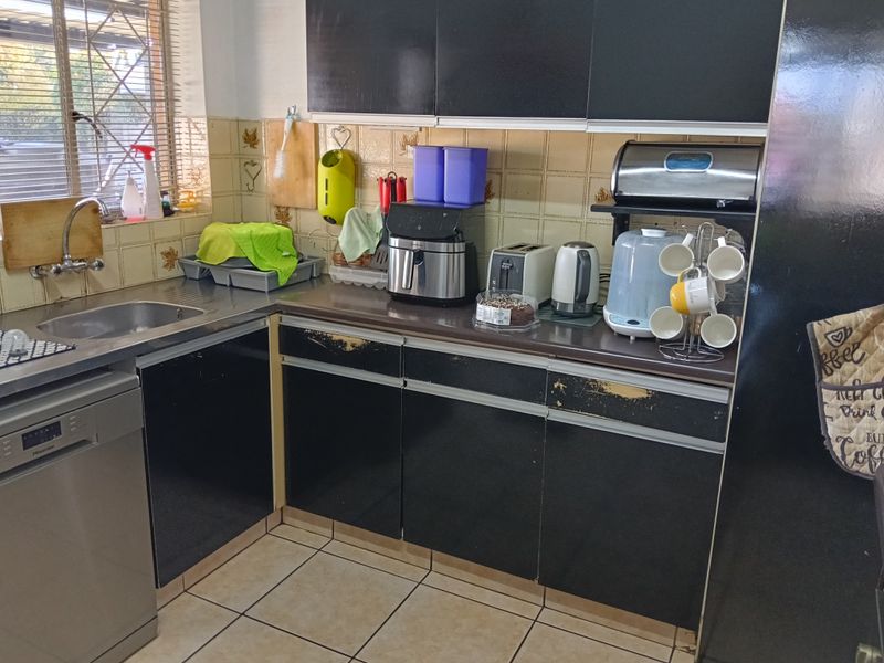 A functional kitchen featuring dark cabinetry with visible wear, stainless steel sink, integrated dishwasher, tiled backsplash, and ceramic floor tiles.