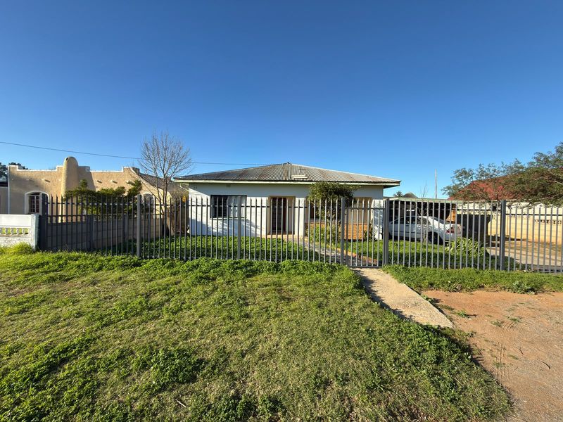 Front view of a single-story house featuring a corrugated metal roof, metal perimeter fencing, and a concrete pathway leading to the entrance.