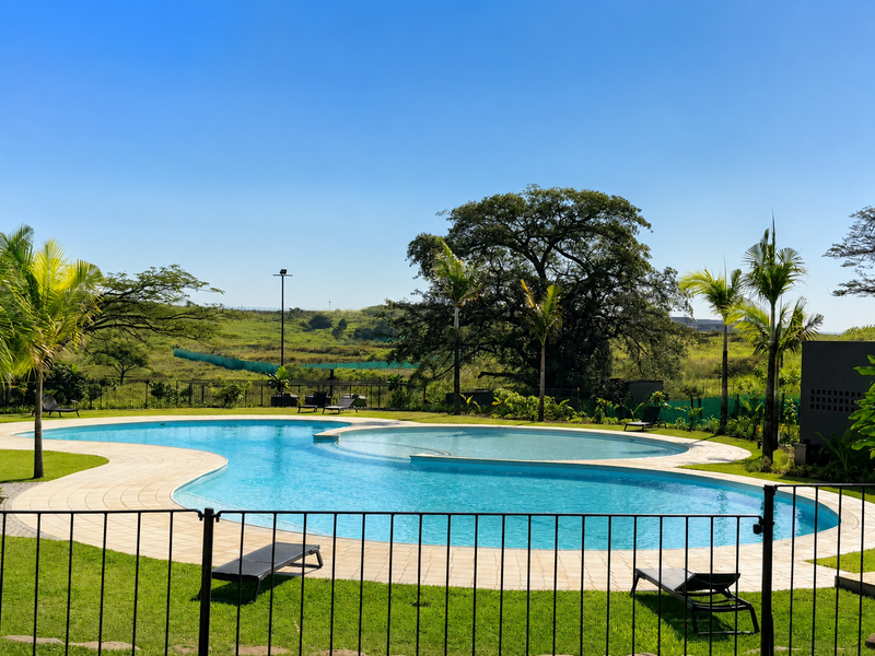 A well-maintained outdoor swimming pool area featuring a curved design, paved surround, and secure metal perimeter fencing set against a scenic landscape.