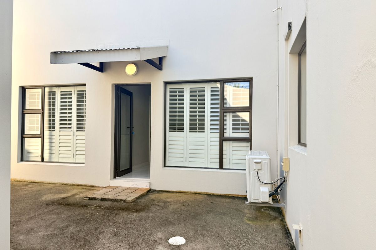 Rear exterior view of the property featuring white walls, aluminium windows with plantation shutters, a metal awning, and an external air conditioning unit.