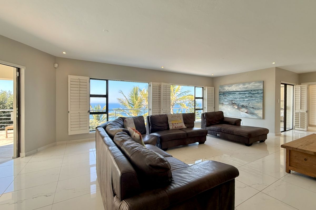Spacious living area featuring polished tiled flooring, recessed lighting, and large windows with plantation shutters offering scenic ocean views and access to a balcony.
