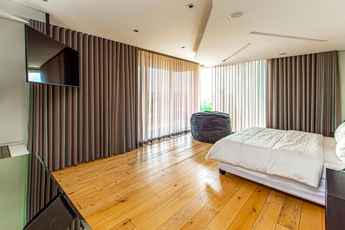Spacious bedroom featuring warm hardwood flooring, a modern dropped ceiling with recessed lighting, and floor-to-ceiling curtains providing ample natural light.