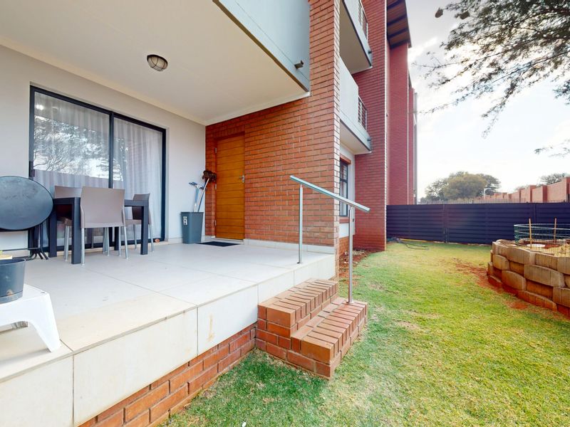 A covered patio area featuring tiled flooring, brick accents, and direct access to the garden via brick steps with a metal handrail.
