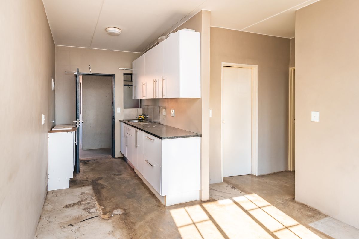 A compact, empty kitchen area featuring white built-in cabinetry, stone countertops, and a stainless steel sink, set against unfinished walls and concrete flooring.