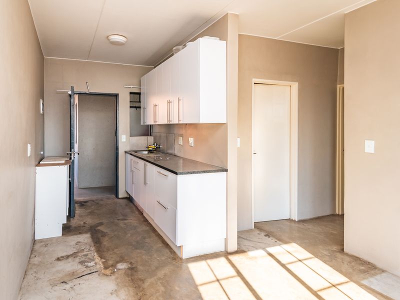A compact, empty kitchen area featuring white built-in cabinetry, stone countertops, and a stainless steel sink, set against unfinished walls and concrete flooring.