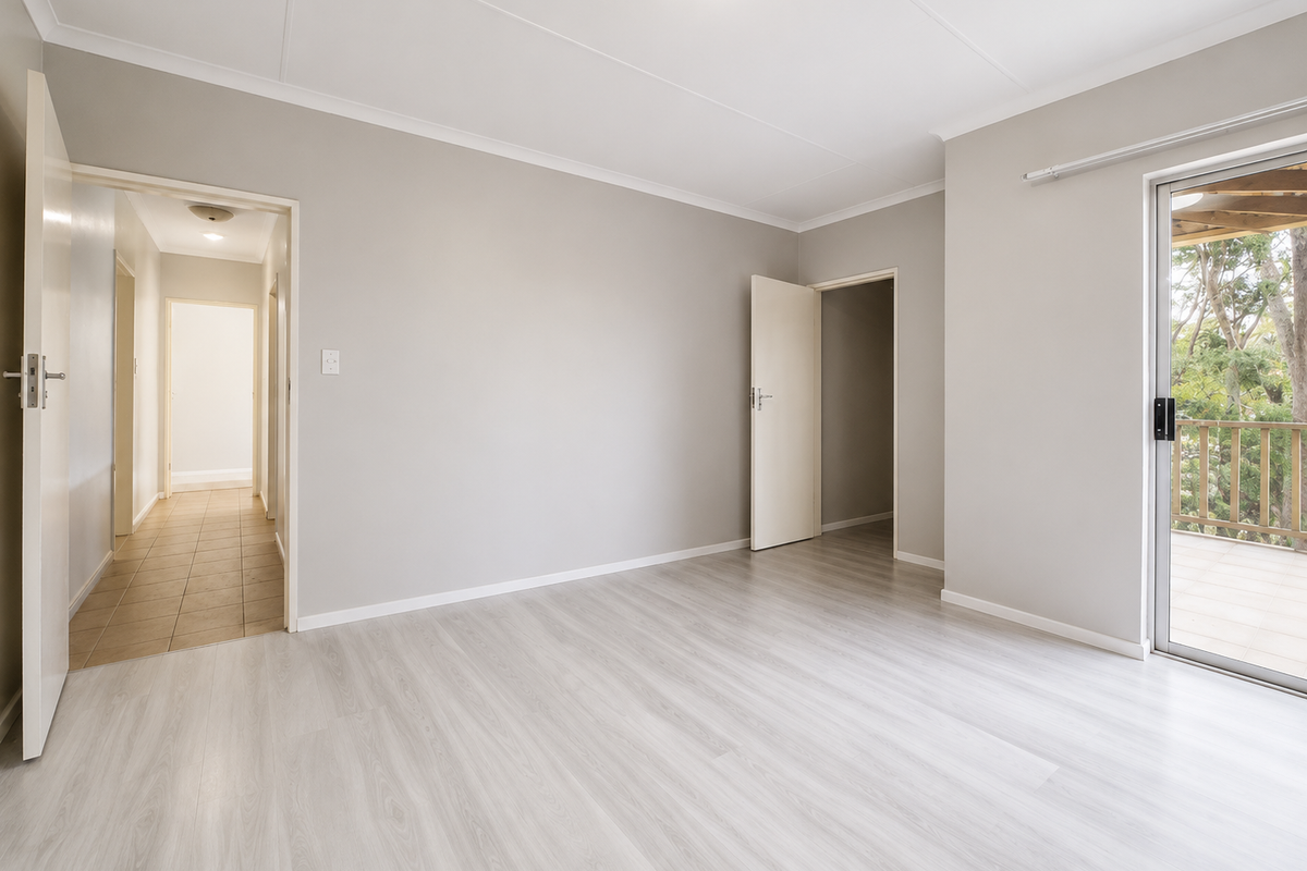 A bright, empty bedroom featuring light-toned laminate flooring, neutral walls, and a sliding glass door providing access to an outdoor balcony area.