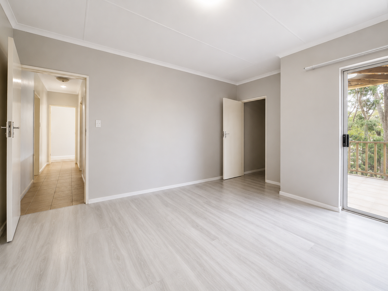 A bright, empty bedroom featuring light-toned laminate flooring, neutral walls, and a sliding glass door providing access to an outdoor balcony area.