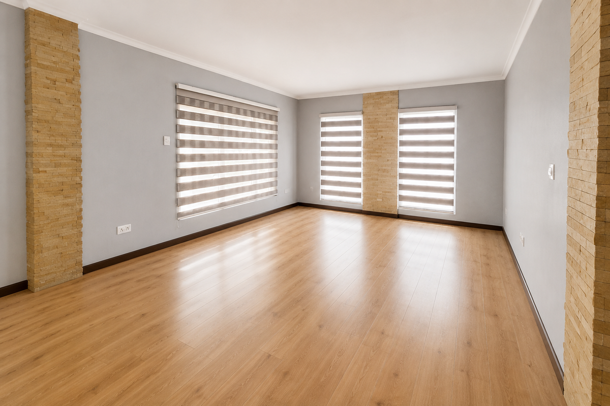 A bright, empty living room featuring light wood-look laminate flooring, neutral grey walls, and decorative stone veneer columns between windows with zebra blinds.