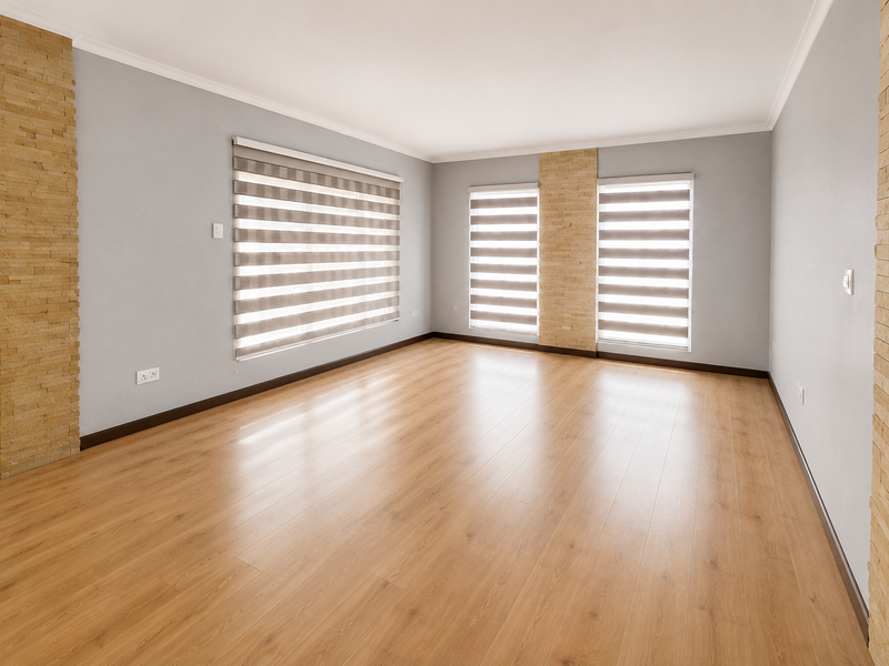 A bright, empty living room featuring light wood-look laminate flooring, neutral grey walls, and decorative stone veneer columns between windows with zebra blinds.