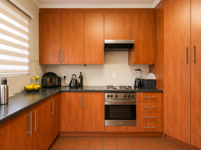 A well-maintained kitchen featuring warm-toned wooden cabinetry, dark laminate countertops, built-in oven with hob, and durable tiled flooring.
