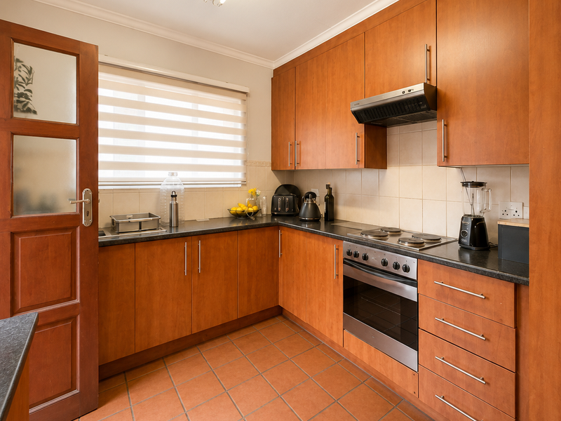 A functional kitchen featuring warm-toned wooden cabinetry, dark laminate countertops, an integrated oven and hob, and durable terracotta-style floor tiles.