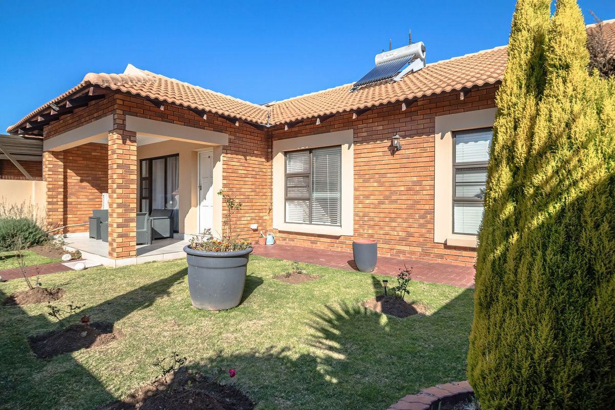 Rear view of a face brick cluster home featuring a covered patio, landscaped garden, and solar water heating system on a tiled roof.