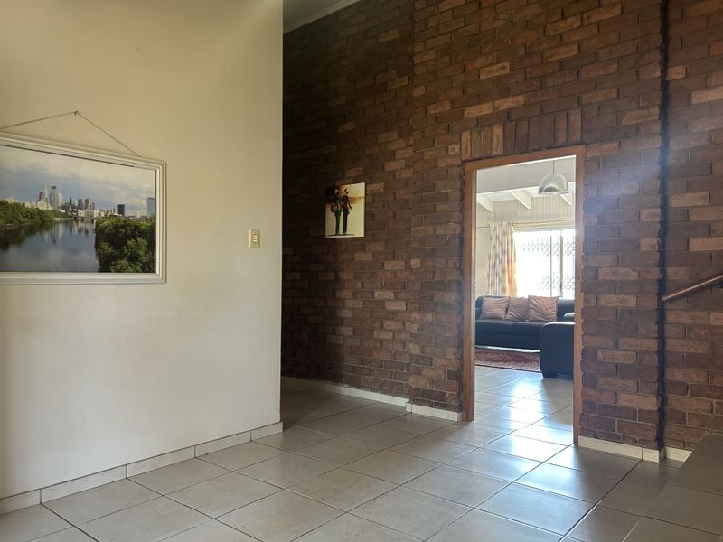 A spacious hallway featuring a prominent exposed brick feature wall and neutral ceramic floor tiles, leading through to an adjacent living area.