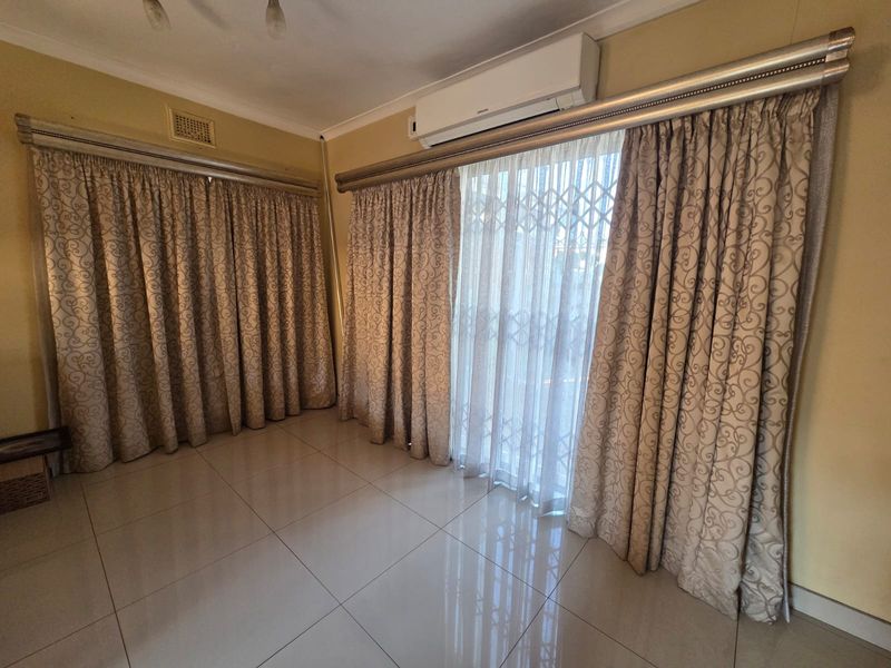 A bright, empty living room featuring large windows with curtains, tiled flooring, and a wall-mounted split-system air conditioning unit.