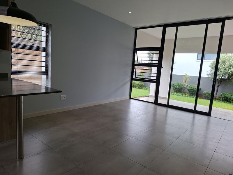 A bright, open-plan living area featuring large sliding glass doors that open to a garden, tiled floors, and a modern kitchen breakfast bar.