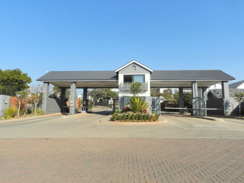 The main entrance and security gatehouse of the townhouse complex, featuring a paved driveway, boom gates, and well-maintained landscaping.