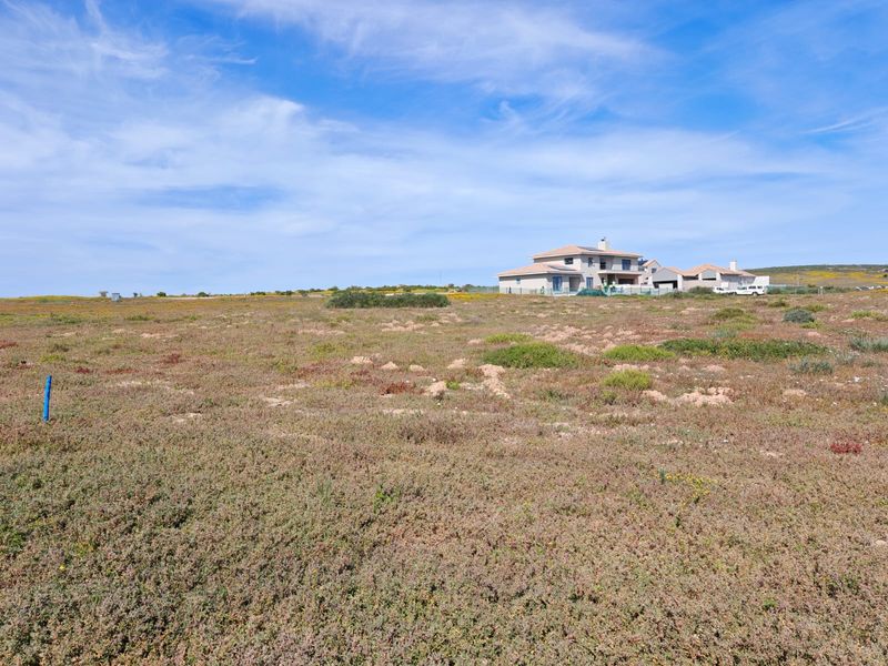 A view of a vacant residential plot of land with natural vegetation and a survey marker, showing surrounding residential development in the distance.