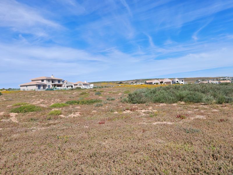 A wide-angle view of a vacant residential plot featuring natural coastal vegetation with established residential homes visible in the background.