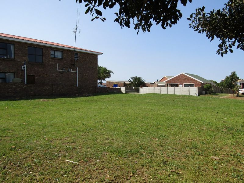 A vacant residential plot featuring a level grassed area, enclosed by a precast concrete boundary wall and adjacent to existing brick residential structures.